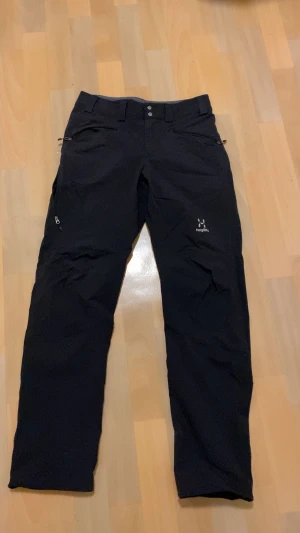 Haglöfs Gabbro täckbyxor  - Svarta Haglöfs Gabbro täckbyxor i storlek S. Byxorna har flera dragkedjeförsedda fickor, bälteshällor och en rak passform. Perfekta för utomhusaktiviteter och gjorda i slitstarkt syntetmaterial. Snyggt Haglöfs-tryck på benet.