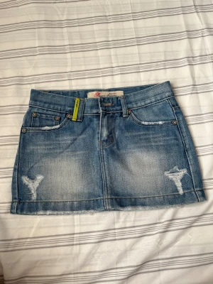 Blå jeanskjol från Paragon  Industries - Snygg blå jeanskjol från Paragom Industries med slitna detaljer och rå kant nedtill. Klassisk femficksmodell med bälteshällor och ljus tvätt. Perfekt för dig som gillar en avslappnad och cool stil.