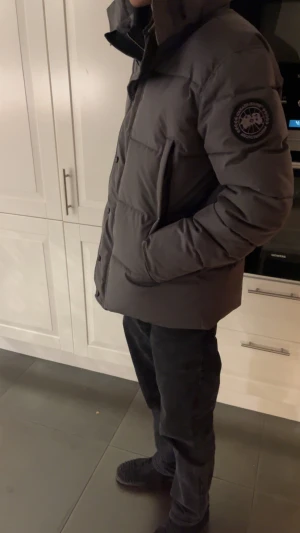 Grå dunjacka från Canada Goose - Säljer en grå dunjacka från Canada Goose med stor huva och logotyp på ärmen. Jackan har en quiltad design, knappar och dragkedja framtill samt två stora fickor. Perfekt för kalla dagar och har en clean, modern look. Fick den som en present men har inte kommit till någon användning 