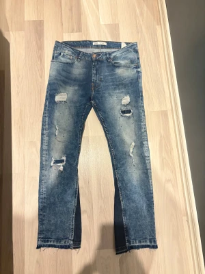 Blå slitna jeans från Zara fleard - Säljer ett par blå jeans från Zara med slitningar och hål på benen för en riktigt edgy look. Jeansen har råa, fransiga benslut och klassisk femficksdesign. Perfekta för dig som gillar streetstyle och vill ha ett par jeans med attityd.