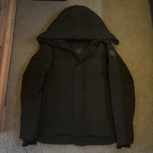 Svart dunjacka från Canada Goose - Svart dunjacka från Canada Goose med huva och klassisk patch på ärmen. Jackan har dragkedja, långa ärmar och en clean, quiltad design. Perfekt för kalla dagar och har en snygg, enkel look som funkar till det mesta.