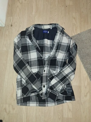 Rutig overshirt från Jack & Jones - Snygg overshirt från Jack & Jones i svart, vit och grått rutigt mönster. Modellen har långa ärmar, knappar framtill och två stora fickor med lock. Perfekt att slänga över en t-shirt för en chill look. Materialet är mjukt och känns som bomull. Hör av er vid funderingar eller prisförslag🙌🏼