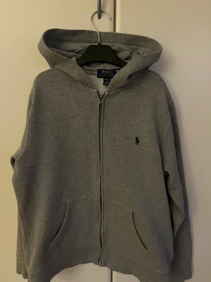 Grå hoodie från Polo Ralph Lauren - Snygg grå hoodie från Polo Ralph Lauren med dragkedja och klassisk huva. Tröjan har två fickor framtill och den ikoniska Polo-loggan broderad på bröstet. Tröjan är i storlek 14/16 som motsvarar xs i vuxen storlek och L i barnstorlek, priset är inte hugget i sten!