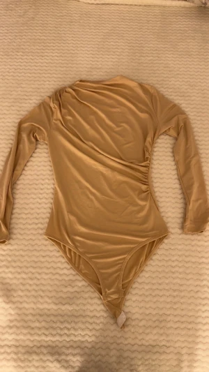 Beige omlott body med lång ärm - Snygg beige body med omlott-effekt och rynkade detaljer vid axel och midja. Långärmad och tillverkad i mjukt, stretchigt syntetmaterial. Praktisk knäppning i grenen med vita tryckknappar. Perfekt för dig som gillar stilrena och enkla plagg med en twist. ALDRIG ANVÄNTS!!