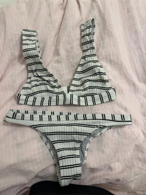 Vit och svart randig bikini - Snygg bikini med ribbad struktur i vitt med svarta ränder. Toppen har breda axelband och en klassisk knäppning bak, medan bikinitrosan har låg midja och smalare sidor. 