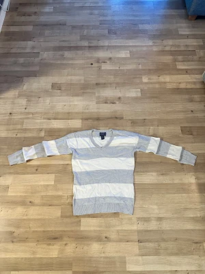 Randig v-ringad tröja från Chaps storlek S - Ljusgrå och vit v-ringad tröja från Chaps med breda ränder. Tröjan har långa ärmar och ribbade muddar vid ärmslut och nederkant. Tillverkad i en mjuk blandning av bomull och modal för en skön känsla.