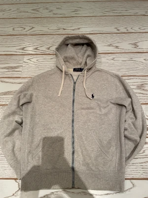Grå hoodie från Polo Ralph Lauren XS - Snygg grå hoodie från Polo Ralph Lauren i storlek XS. Tröjan har huva med snörning, dragkedja framtill och två fickor. Klassisk Polo-logga broderad på bröstet. Mjuk bomullskvalitet och ribbade muddar vid ärmslut och nederkant.
