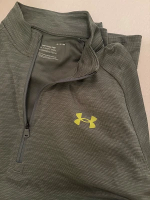 Grön half zip träningströja Under Armour - Grön långärmad träningströja från Under Armour med diskret mönster och gul logga på bröstet. Tröjan har half zip framtill och är gjord i lätt polyester med skön passform, perfekt för sport och aktiva dagar.