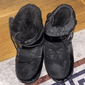 Svarta moon boots med snörning - Säljer ett par svarta moon boots med glansiga detaljer och snörning runt skaftet. Insidan är fodrad med mjukt syntetmaterial för extra värme. Yttermaterialet är syntet och sulan är platt med mönster för bra grepp. Perfekta för kalla vinterdagar.