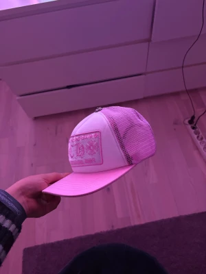 Chrome hearts - Snygg truckerkeps i vitt och ljusrosa med mesh på sidorna och bak. Framtill finns en broderad patch med CH-logga och blommor i rosa. Kepsen har böjd skärm och metallknopp på toppen. Perfekt för dig som gillar streetstyle och vill sticka ut.