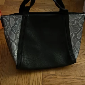 Svart handväska med ormmönster - Snygg svart handväska med detaljer i grått ormmönster på sidorna. Väskan har två rejäla handtag, dragkedja upptill och ett extra fack med dragkedja på baksidan. Insidan är ljus och har flera fack för smart förvaring. Perfekt för dig som vill sticka ut lite extra.