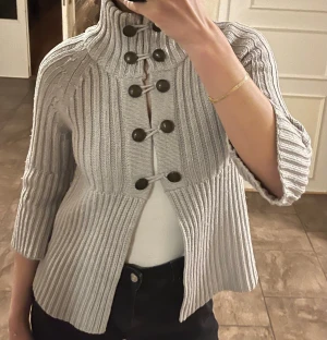 Grå stickad kofta med trekvartsärmar - Säljer en snygg grå ribbstickad kofta med hög krage och dekorativa stora knappar framtill. Koftan har trekvartsärm. Storlek XS, men passar bra på mig som vanligtvis bär S. 