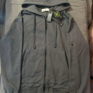 Mörkgrå hoodie från Stone Island - Snygg mörkgrå hoodie från Stone Island med klassisk logopatch på ärmen. Tröjan har huva med snörning, två stora fickor fram och dragkedja hela vägen. Tillverkad i mjuk bomull och har ribbade muddar. Perfekt för dig som gillar streetwear och vill ha något stilrent.