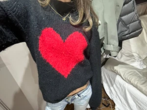 Svart stickad tröja med rött hjärta - Mysig svart stickad tröja med ett stort rött hjärta på bröstet. Tröjan har lång ärm och en högre hals, perfekt för att ge en cool och gullig vibe. Materialet är fluffigt och mjukt, vilket gör den extra skön att bära. Passar dig som vill sticka ut med en söt detalj. Står inget märke, Jag köpte den i London förra vintern och använt fåtal gånger