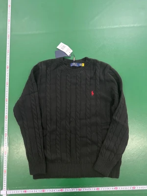 Svart kabelstickad tröja Polo Ralph Lauren - Svart kabelstickad tröja från Polo Ralph Lauren i storlek XL. Klassisk rund hals och långärmad modell med ribbade muddar. Tröjan har den ikoniska röda Polo-loggan broderad på bröstet och är tillverkad i mjuk bomull. Perfekt för dig som gillar stilren och tidlös design.