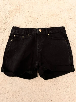 Svarta denimshorts med uppvik - Snygga svarta denimshorts med klassisk femficksdesign och uppvikta benslut. Shortsen har guldiga nitar och knapp, samt bälteshällor i midjan. Perfekta för en chill och stilren look under varma dagar.