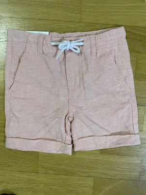 Ljusrosa shorts med snörning, strl 92 - Snygga ljusrosa shorts från C i storlek 92. De har en vit snörning i midjan, uppvikta benslut och praktiska fickor både fram och bak med knappdetaljer. Materialet känns lätt och somrigt, perfekt för varma dagar.