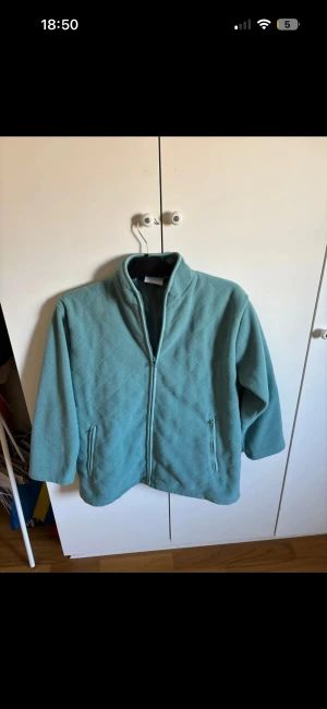 Turkos fleecejacka från Genevan - Turkos fleecejacka från Genevan i storlek M. Jackan har ett quiltat mönster, dragkedja framtill och två sidofickor med dragkedja. Hög krage och långärmad modell, perfekt för kyliga dagar. Enkel och stilren design som är lätt att matcha.