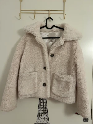 Beige teddyjacka med stora fickor från AMISU - Mysig teddyjacka i beige färg med fluffigt material och stora framfickor i storlek 34 från AMISU. Jackan har bred krage, tre knappar framtill och långärmade ärmar. 