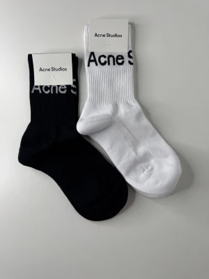 Svarta och vita Acne Studios strumpor - Två par ribbade strumpor från Acne Studios, ett par svarta och ett par vita, båda med logotyp upptill. Tillverkade i mjuk bomullsblandning med 90% bomull, 8% nylon och 2% elastan. Perfekta för en clean och stilren look.