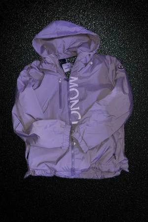 Grå vindjacka från Moncler - Snygg grå vindjacka från Moncler med huva och dragkedja framtill. Jackan har Moncler-logga på ärmen och stor vit Moncler-text längs bröstet. Tillverkad i polyester, med elastiska muddar och praktiska fickor. Perfekt för dig som gillar stilren och sportig look.