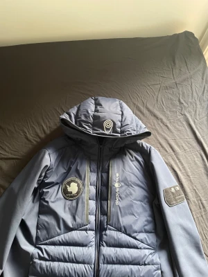 Blå Sail Racing dunjacka med huva - Snygg mörkblå dunjacka från Sail Racing med huva och coola patchar på bröstet och ärmen. Jackan har dragkedja framtill, quiltad design och logga på insidan. Perfekt för dig som gillar sportig stil och vill hålla dig varm under kalla dagar.