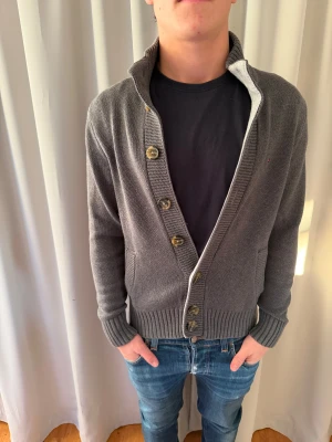 Grå kofta från Tommy Hilfiger XS - Snygg grå kofta från Tommy Hilfiger i stickad bomull. Modellen har ribbad krage, muddar och nederkant, samt knappar framtill. Liten broderad logga på bröstet och två fickor. Perfekt för dig som gillar klassisk stil med en modern touch.