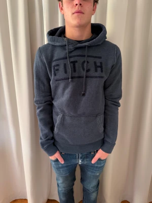 Mörkgrå hoodie Abercrombie & Fitch - Säljer en mörkgrå hoodie från Abercrombie & Fitch i storlek M. Tröjan har broderad FITCH-logga på bröstet, snörning i huvan och en stor magficka. Mjuk insida och klassisk passform, perfekt för chill dagar. Materialet är bomull och polyester.