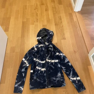 Blåvit barn hoodie från Burton med huva - Snygg vindjacka från Burton i blått och vitt batikmönster. Jackan har huva, hel dragkedja och är tillverkad i mjukt syntetmaterial med fleecefoder på insidan. Perfekt för dig som gillar streetstyle och vill sticka ut lite extra. Barn Hoddie med storlek XS.