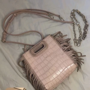 rosa axelväska med fransar från Maje - Säljer en rosa axelväska från Maje med croco-mönstrad yta och snygga fransar längs sidorna. Väskan har både axelrem och en chunky kedja, samt ett praktiskt handtag upptill. Perfekt för dig som vill sticka ut med en cool accessoar.