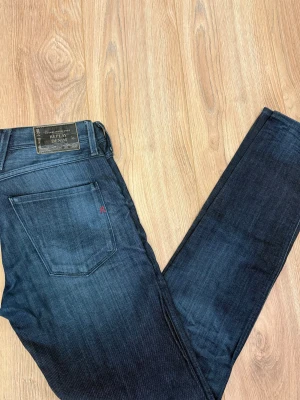 Replay Anbass jeans mörkblå 30/32 - Snygga Replay Anbass jeans i mörkblå tvätt med klassisk femficksdesign och diskret Replay-logga på bakfickan. Jeansen har normal passform och raka ben, tillverkade i slitstarkt bomullsmaterial. Perfekt för dig som gillar stilrena och tidlösa jeans.