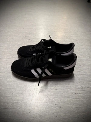 Adidas Spezial svarta sneakers - Svarta Adidas Spezial sneakers med klassiska vita ränder på sidorna och guldfärgad Spezial-logga. Skorna har snörning, platt sula och är tillverkade i mocka med detaljer i syntet. Rund tå och låg profil ger en clean och sportig vibe.