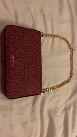 Vin Röd axelväska från Michael Kors - Snygg vin röd  axelväska från Michael Kors med guldfärgad kedja som axelrem. Väskan har ett geometriskt mönster med MK-loggor och är tillverkad i syntetmaterial. Perfekt storlek för det viktigaste och har stilren design med logotyp i guld på framsidan.