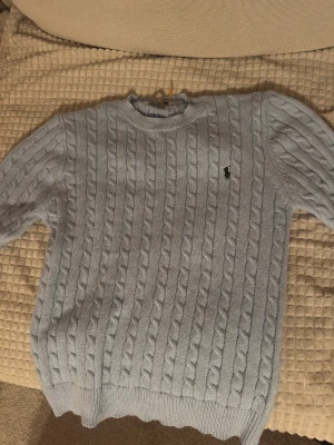Ljusblå stickad tröja Polo Ralph Lauren - Säljer en ljusblå stickad tröja från Polo Ralph Lauren med klassiskt kabelstickat mönster och rund hals. Tröjan har långärmade ärmar och den ikoniska Polo-loggan broderad på bröstet. Perfekt för dig som gillar stilren och tidlös design.