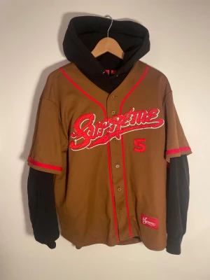 Supreme brun baseball tröja med huva - Supreme baseball tröja i brun färg med röda detaljer och broderad logga framtill. Nummer 5 i rött på både fram- och baksida. Kortärmad design med svart huva och svarta långa ärmar under. Materialet är bomull/polyesterblandning. Snygg streetwear-vibe och knappar framtill.