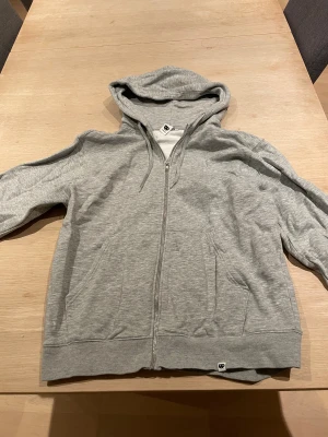 Grå hoodie med dragkedja från Junkyard - Snygg grå hoodie från Junkyard i storlek M. Tröjan har huva med snörning, praktiska fickor fram och hel dragkedja. Tillverkad i mjuk bomullsmix som är skön att ha på sig. Perfekt för chill dagar eller när du vill ha en enkel och clean look. Använd några gånger men inga skador på tröjan.