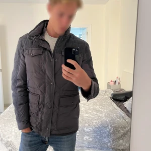 Grå quiltad pufferjacka med ullpaneler - Säljer en grå pufferjacka med quiltad design och snygga ullpaneler på axlar och ärmar. Jackan har fyra fickor framtill med lock och knappar, hög krage och stängs med både dragkedja och knappar. Perfekt för dig som vill ha en stilren och varm jacka med coola detaljer.