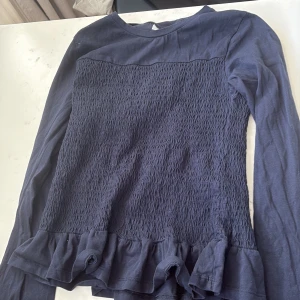 Mörkblå långärmad topp Gina Tricot - Supersöt mörkblå långärmad topp från Gina Tricot med smockad framsida och volangkant nedtill. Perfekt för dig som gillar en lite sötare stil. Mjuk och skön i bomullsmaterial.