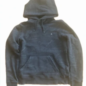 Svart hoodie från J.Lindeberg - Snygg svart hoodie från J.Lindeberg med diskret logomönster över hela tröjan. Tröjan har huva med snörning, magficka och långa ärmar. Perfekt för dig som gillar stilrena detaljer och sportig look. Tveka inte på att höra av er vid frågor!