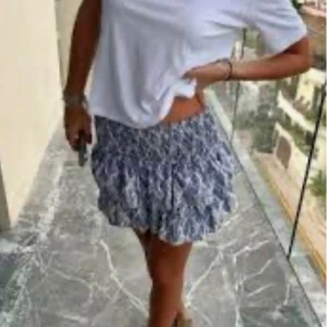 Vit och blå kortkjol med volanger  - Kortkjol med blått och vitt mönster, resår i midjan och lätta volanger. Meet Me There, storlek M, nytt skick. Nypris 398 kr – säljes för 210 kr! 🩵🩵