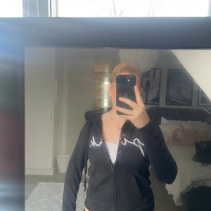 Svart hoodie från Victoria’s Secret med dragkedja - Svart hoodie från Pink med dragkedja framtill och huva. Tröjan har ett stort tryck i vitt och pastellfärger över bröstet. Mjuk och skön, perfekt för chill dagar. Klassisk passform med långa ärmar och fickor framtill.