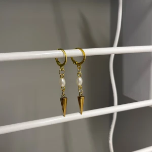 Guldiga örhängen med pärlor och spikes - Unika örhängen i guldton med små vita pärlor och coola konformade spikes som hänger ner. Perfekt för dig som vill ha något som sticker ut och ger en edgy vibe till din stil. I rostfrittstål och handgjort