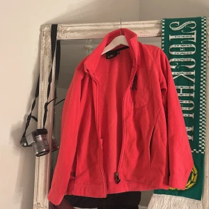 Röd fleecejacka från McKinley - Röd fleecejacka från McKinley med hög krage och hel dragkedja. Jackan har två fickor med dragkedja och en enkel, sportig look. Perfekt för kyliga dagar och outdoor-häng. Mjuk och skön känsla, passar dig som gillar att vara aktiv och om du har storlek 140