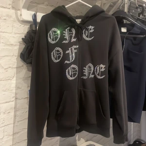 Svart zip hoodie med glitter detaljer från One of One - Svart zip hoodie från One of One i storlek Large, med huva och snörning. Framsidan har coola glitter detaljer som bildar text i ren stil. Mjuk insida, och perfekt för dig som gillar streetwear och vill sticka ut.