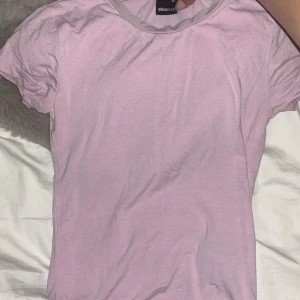 Ljusrosa t-shirt från Gina Tricot - En enkel och mjuk ljusrosa t-shirt från Gina Tricot med rund hals och korta ärmar. T-shirten har en ljusgrå kant vid halsen som ger en snygg kontrast. Perfekt för en chill och avslappnad stil.