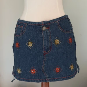 Broderad jeanskjol med snörning - Mörkblå jeanskjol med broderade blommor i rött och gult framtill och dekorativ brodyr på bakfickan. Kjolen har snörning längs ena sidan och röda kontrastsömmar. Klassisk femficksmodell i bomull med knapp och dragkedja framtill.