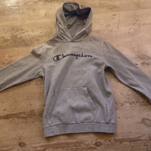 Grå Champion hoodie med huva - Grå hoodie från Champion med svart logga på bröstet. Tröjan har huva, magficka och långa ärmar. Perfekt för chill dagar och streetwear. Materialet är mjukt och känns som bomull och polyester. Passformen är normal och modellen är klassisk.