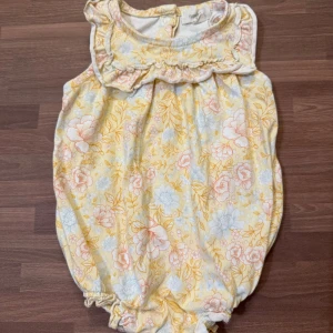 Blommig romper - Använd väldigt fåtal gånger och utan anmärkningar. Supersöt romper från Newbie i stl 62 (2-4mån). Ljusgul med blommigt mönster med volang vid halsen och knappar bak samt tryckknappar i grenen.