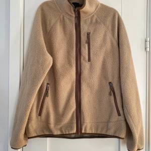 Beige teddyjacka från 157, storlek S - Mysig beige teddyjacka från 157 i storlek S. Jackan har bruna detaljer vid dragkedjorna, hög krage och tre fickor med dragkedja framtill. Tillverkad i fluffigt fleece-material som håller dig varm under kyliga dagar. Jackan har en liten fläck vid ena fickan i övrigt i bra begagnat skick.  Mått Över bröstet: ca 59 cm Längd från axeln och ner: ca 66 cm  100% polyester  Kommer från ett djur och rökfritt hem. 