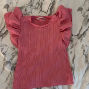 Rosa ribbad topp med volangärmar från Zara - Säljer en rosa ribbad topp från Zara i storlek S. Toppen har korta, voluminösa volangärmar som ger en cool och trendig look. Materialet är mjukt och stretchigt, perfekt för dig som gillar att sticka ut med färg och detaljer.